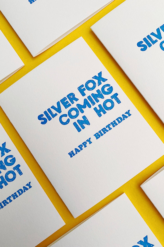 greeting card with 'Silver Fox Coming In Hot' printed in blue ink on the front, accompanied by 'HAPPY BIRTHDAY' in smaller text. The cards are on a yellow background.
