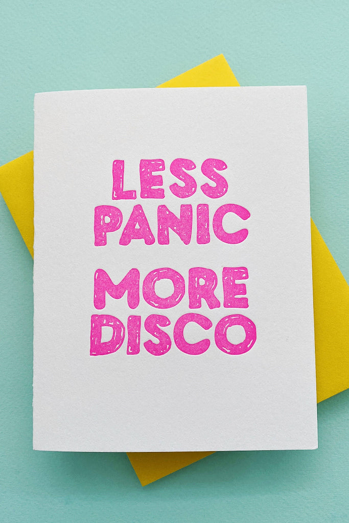 A greeting card with 'LESS PANIC MORE DISCO' printed in pink letters on a white background, accompanied by a yellow envelope.