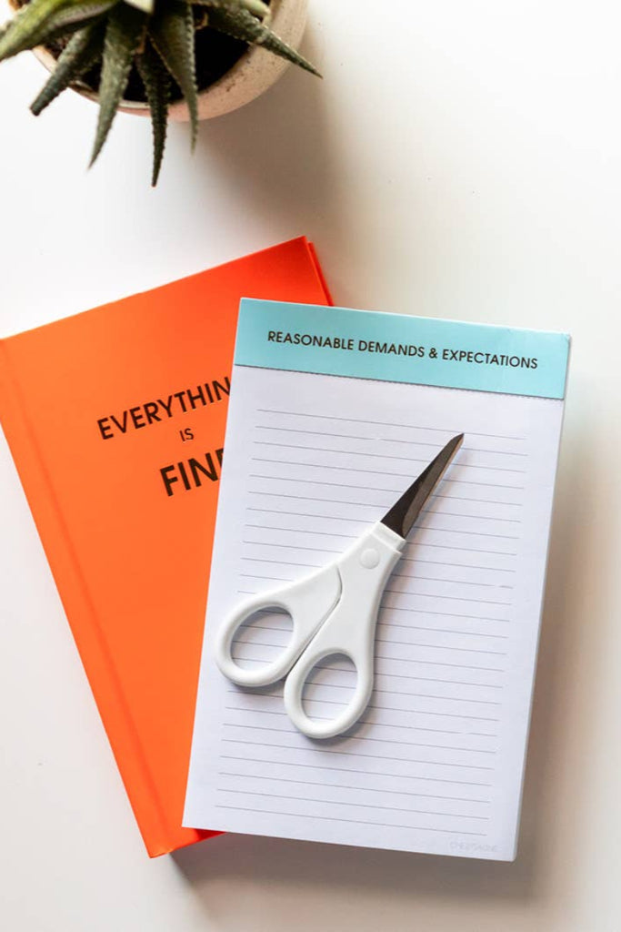 Notebook with scissors on a white surface, next to an orange book and a plant. says reasnable demands & expectations

