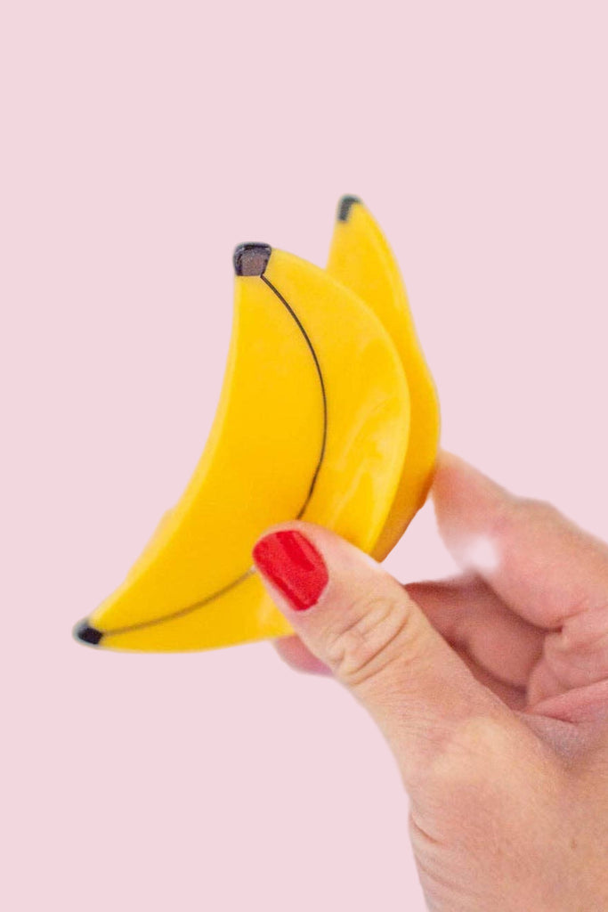 Hand holding a small yellow banana-shaped hair clip against a light pink background