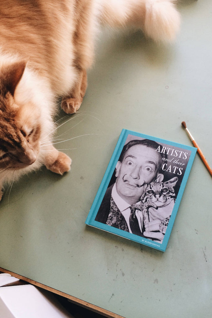 Cat interacting with a book titled 'Artists and their Cats' on a green surface.