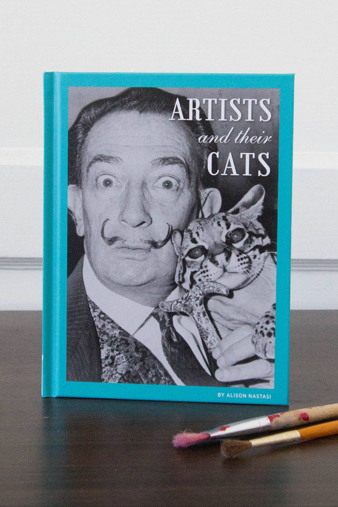 Book titled 'Artists and their Cats' with a cover featuring a famous artist holding a cat, placed on a surface with paintbrushes.
