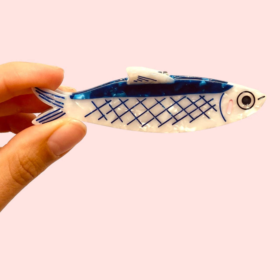 Fish-shaped anchovy hair cliip with blue and white pattern held against a pink background