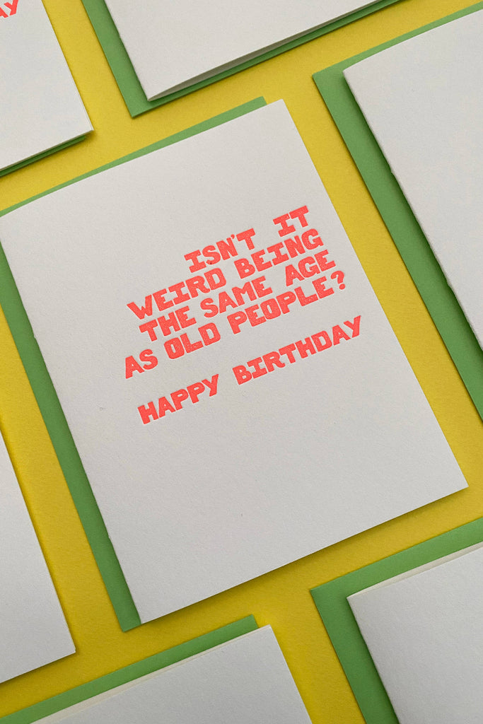 Greeting cards with orange text 'ISN'T IT WEIRD BEING THE SAME AGE AS OLD PEOPLE? HAPPY BIRTHDAY' laid out on a yellow background.