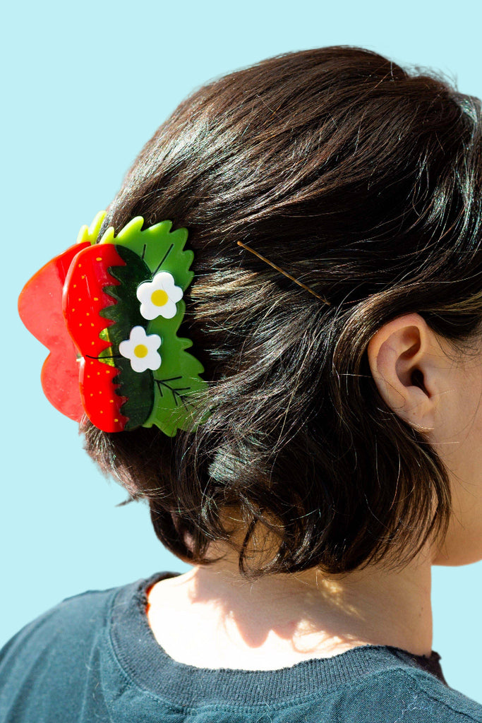 Person with a decorative hair clip featuring red strawberries, white flowers and green leaves against a pale blue background