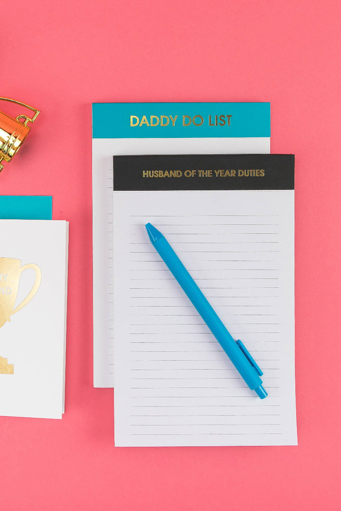 Notebook, pen, and card on a pink background with trophy and bell decorations. husband of the year
