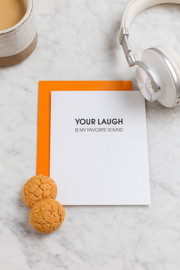 Note with 'Your laugh is my favorite sound' text, headphones, and cookies on a marble surface
