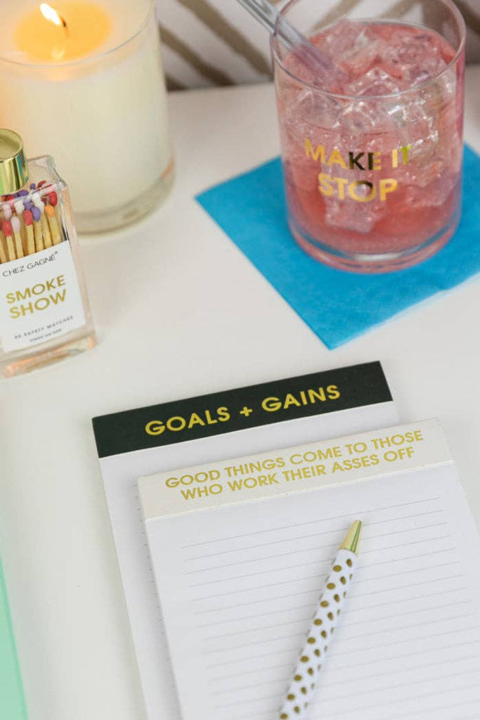 Desk setup with a bottle of 'Smoke Show' matches, a glass with pink liquid and a straw, a notepad with motivational quote, good thingd come to those who work their asses off